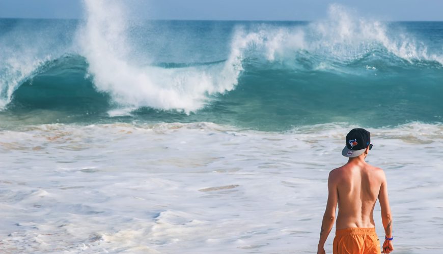 man in orange shorts standing near wavy sea during daytime